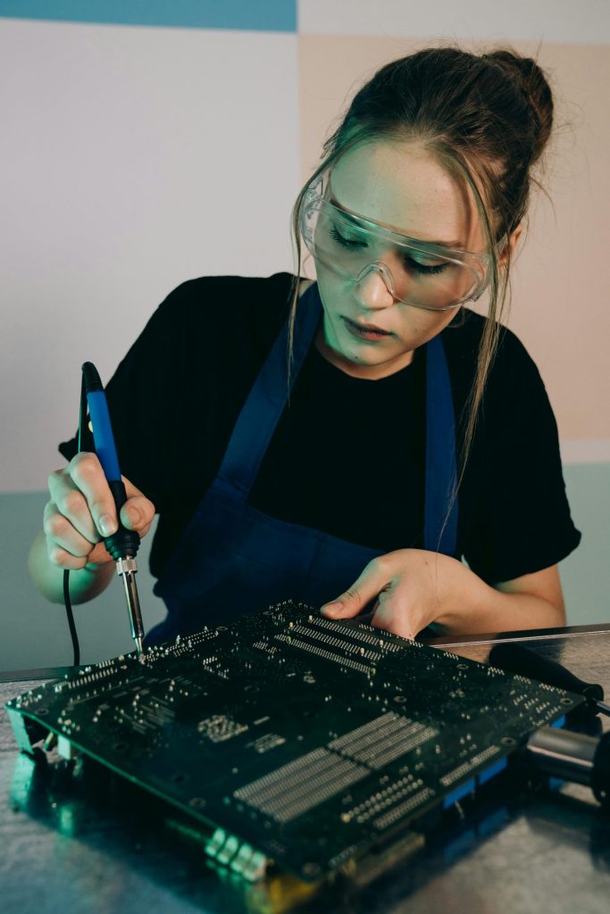 A young woman in an apron and goggles soldering a circuit board indoors.